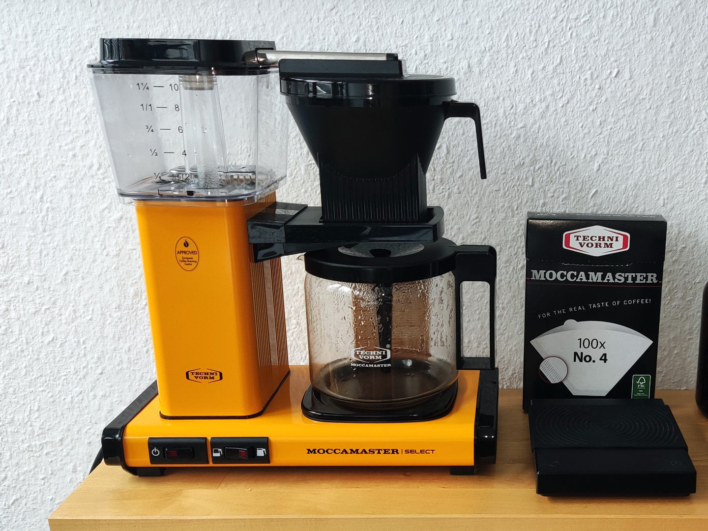 Titelbild zum Blogartikel über die Zubereitung mit der Moccamaster-Kaffeemaschine. Zu sehen ist eine senfgelbe Moccamaster nebst passenden Filtern, einem Kaffee-Pfandglas und einer Kaffeemühle auf einem Holztisch. In weiß ist "Brew Guide" zu lesen.