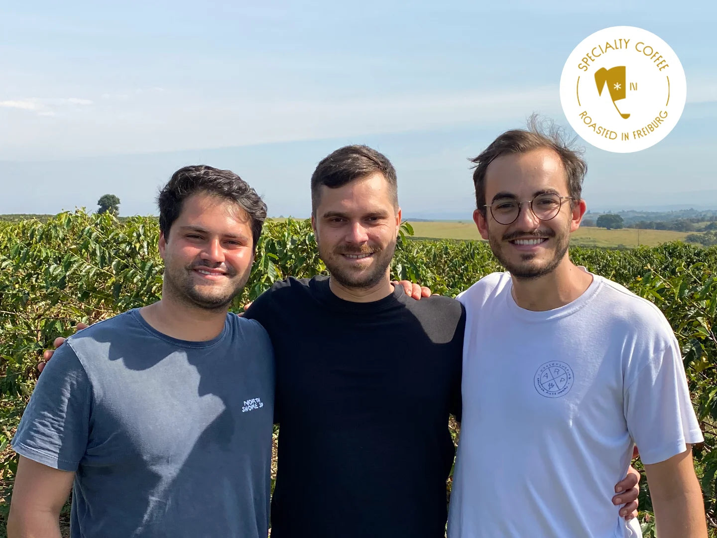 Titelbild zum Blogartikel: Luis, Mats und Niklas (v. l. n. r.) stehen vor Kaffeepflanzen auf einer Farm in Brasilien. Aufgenommen im Juni 2022. In weißer Schrift liest man "Wer ist Ocafi?".