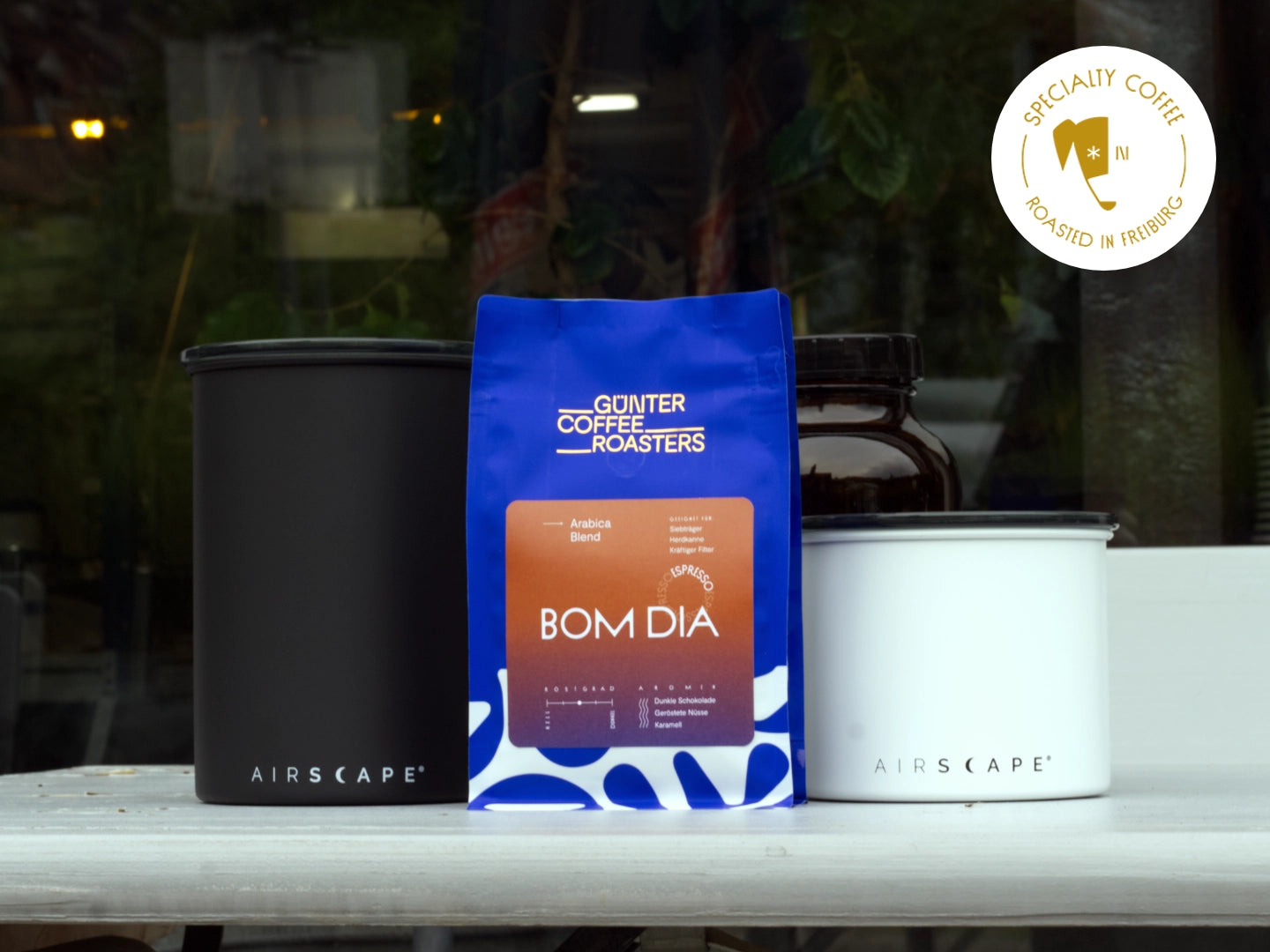 Titelbild zum Artikel über die Kaffeeaufbewahrung. Zu sehen sind eine Verkaufsverpackung der Günter Coffee Roasters in strahlendem Blau mit goldenem Logo und Bom-Dia-Etikett. Links und rechts davon jeweils eine Airscape-Dose in schwarz und weiß.
