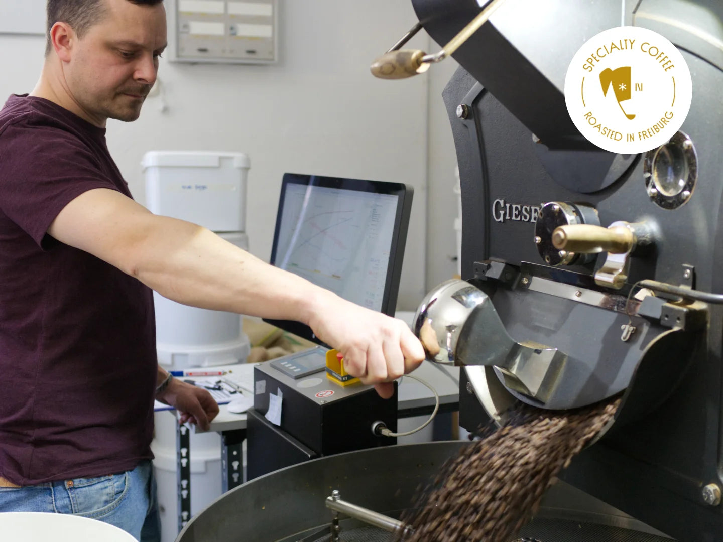 Titelbild zum Blogartikel "Wie wird Kaffee geröstet?". Auf dem Bild ist Philip an der Röstmaschine Giesen W15 zu sehen. Er öffnet gerade die Ablassklappe, um frischen Röstkaffee in die Abkühlschale zu geben. In weiß liest man "Kaffee rösten".
