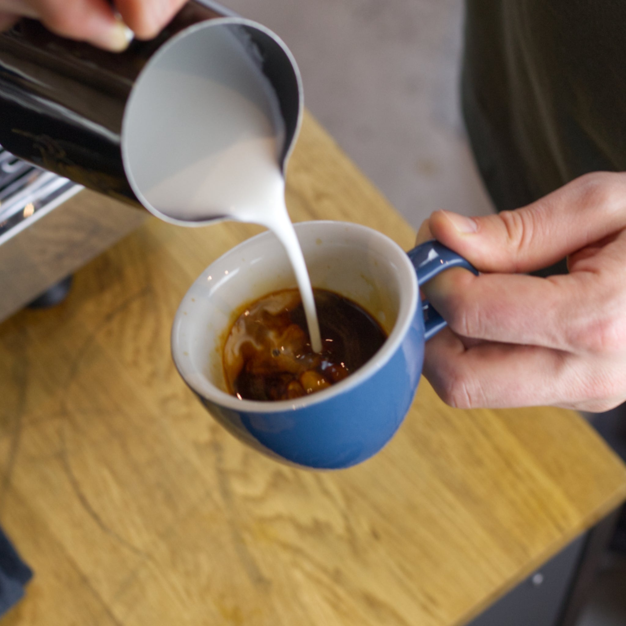 Mats gibt Milch in den Espresso. Dafür verwendet er ein spezielles Milchkännchen, um Latte Art zu gießen.